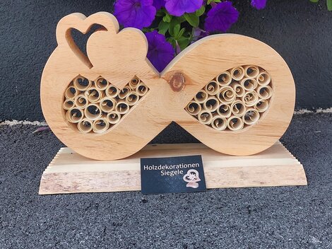 Eine dekorative Holzskulptur in Form eines Unendlichkeitssymbols mit Herzmotiven und einem spiralartigen Muster. Im Hintergrund sind lila Blumen zu sehen.