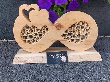 A decorative wooden sculpture in the shape of an infinity symbol with heart motifs and a spiral pattern. In the background, there are purple flowers.