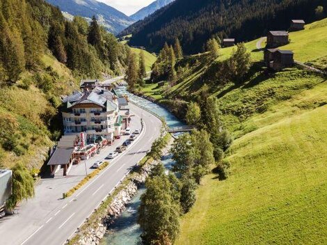 Eine malerische Landschaft mit einem Fluss, der neben einer Straße fließt. Im Hintergrund sind grüne Hügel und einige Chalets zu sehen.
