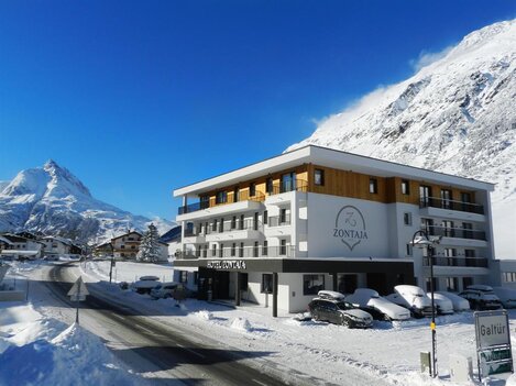 A modern hotel named "Zontaja" in a winter mountain landscape. The surroundings are covered in snow, and majestic mountains can be seen in the background.