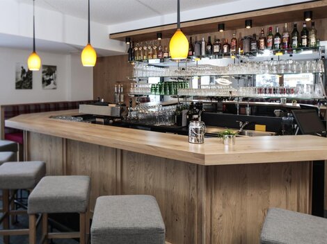 Eine moderne Bar mit einem geschwungenen Holztresen und stilvollen Hockern. Im Hintergrund sind Regale mit einer Auswahl an Spirituosen und Gläsern zu sehen.