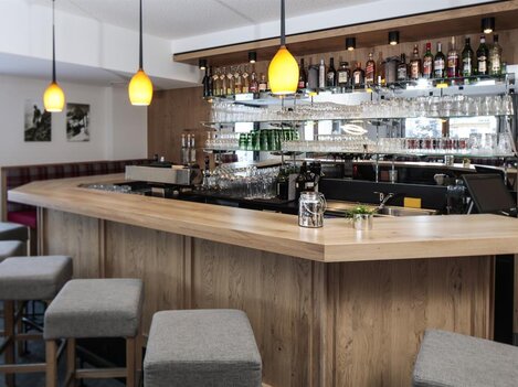 A modern bar with a curved wooden counter and stylish stools. In the background, shelves with a selection of spirits and glasses can be seen.