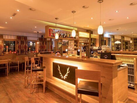 An inviting bar with wooden furniture and dim lighting. In the background, counters and shelves stocked with drinks can be seen.