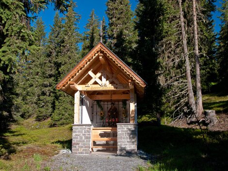 Eine kleine Holzkapelle umgeben von hohen Bäumen. Der Himmel ist blau und die Umgebung ist grün und friedlich. | © Bernhard Gruber – Photography