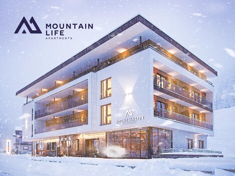 A modern apartment building in the snow, surrounded by a winter landscape. The facade is in light tones and features large windows.