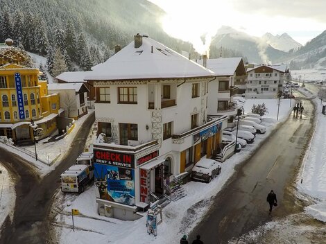 Ein charmantes Gebäude im Schnee mit einer "CHECK IN"-Anzeige. Im Hintergrund sind verschneite Berge und eine ruhige Straße zu sehen.