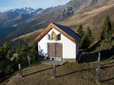 Eine kleine Kapelle auf einem Hügel, umgeben von Bergen und Wäldern. Die Landschaft zeigt klare Himmel und Berggipfel im Hintergrund. | © Bernhard Gruber – Photography