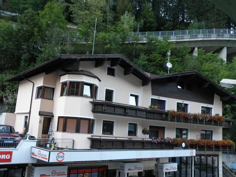 Ein charmantes Gebäude mit mehreren Balkonen und blühenden Fenstern. Im Hintergrund sind Bäume und eine Straßenbrücke sichtbar.