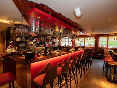 A modern bar with an inviting use of red light. The counter is well-stocked and there are many chairs waiting for guests.