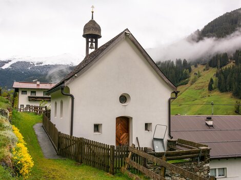 Eine kleine, weiße Kirche mit einem Glockenturm steht in einer malerischen Landschaft. Im Hintergrund sind grüne Hügel und bewölkter Himmel sichtbar. | © Bernhard Gruber – Photography