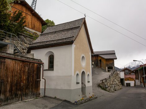 Eine kleine, weiße Kapelle steht an einer engen Straße, umgeben von traditionellen Holzhäusern. Der Himmel ist bewölkt und es scheint eine ruhige, ländliche Umgebung zu sein. | © Bernhard Gruber – Photography