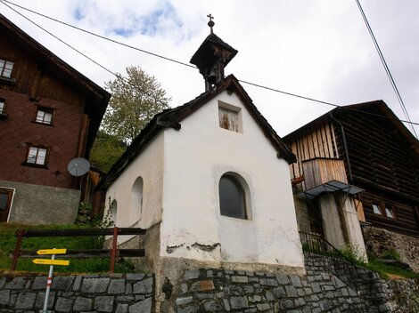 Eine kleine weiße Kapelle zwischen traditionellen Holzhäusern. Im Hintergrund sind Bäume und ein bewölkter Himmel zu sehen.