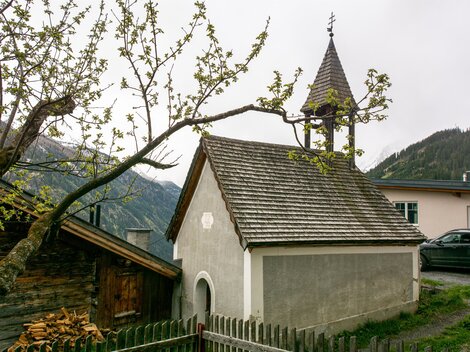Eine kleine Kapelle mit einem spitzen Dach und einem Kirchturm ist umgeben von Bäumen und Bergen. Im Vordergrund steht ein Holzzaun. | © Bernhard Gruber – Photography
