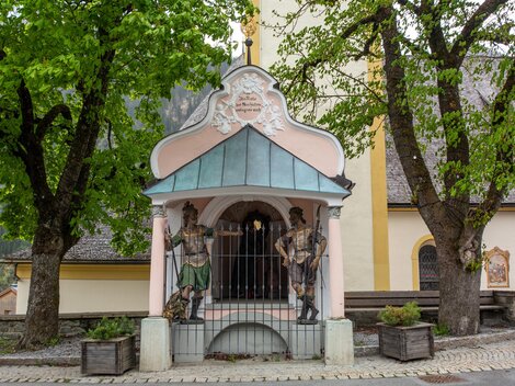 Eine kleine Kapelle mit bunten Figuren und einem grünen Dach. Im Hintergrund stehen große Bäume und ein Gebäude mit gelber Fassade. | © Bernhard Gruber – Photography