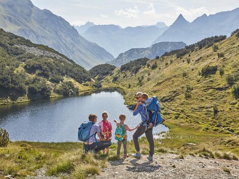 Eine Familie wandert in den Bergen neben einem klaren See. Die Landschaft ist grün und hügelig, umgeben von beeindruckenden Gipfeln.