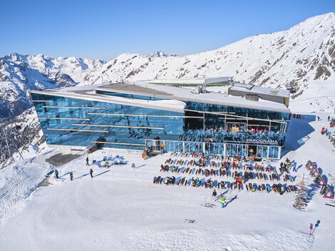 Eine moderne Berghütte umgeben von schneebedeckten Bergen. Viele Menschen genießen den sonnigen Tag im Freien.