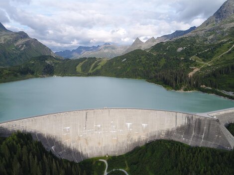 Ein beeindruckender Staudamm umgeben von grünen Bergen und einem klaren See. Die Szenerie zeigt eine ruhige Naturkulisse mit leicht bewölktem Himmel.