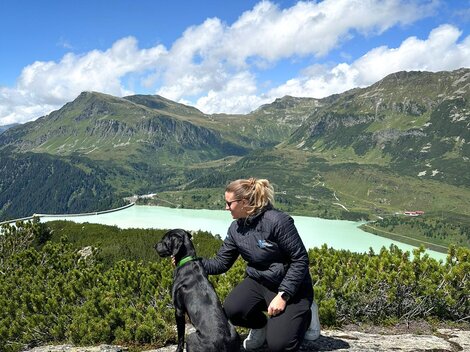 Eine Frau sitzt mit ihrem Hund auf einem Felsen und blickt auf eine grüne Landschaft mit Bergen und einem See. Der Himmel ist blau mit einigen Wolken.