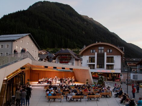 Eine Veranstaltungen im Freien mit einem Orchester spielt vor einer Kulisse aus Bergen. Viele Menschen sitzen auf Bänken und genießen die Musik.