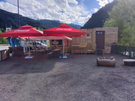 Ein kleiner Imbissstand mit Holzstruktur und roten Sonnenschirmen. Die Umgebung ist bergig und grün, ideal für eine Pause im Freien.