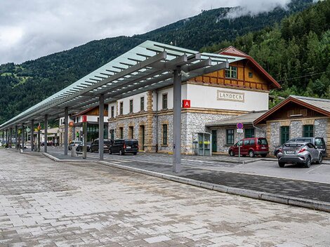 Ein Bahnhofsgebäude mit traditioneller Architektur und moderner Überdachung. Im Hintergrund sind grüne Berge und Wolken sichtbar.