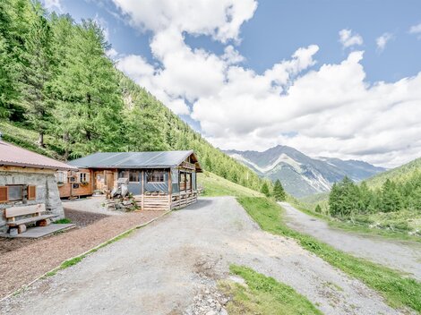 Eine malerische Alpenlandschaft mit zwei Hütten und grünen Bäumen. Die Berge im Hintergrund sind unter einem klaren Himmel sichtbar.