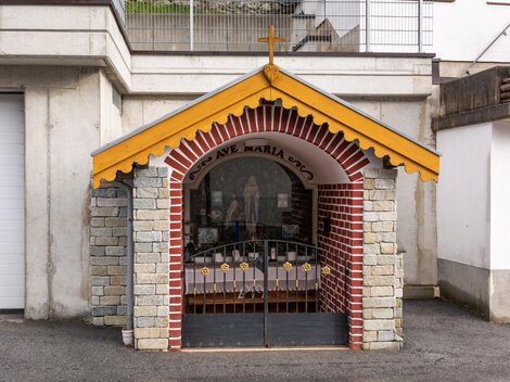 Eine kleine Kapelle mit einem gelben Dach und einem Kreuz. Sie ist aus Stein und Ziegeln gebaut und hat eine geschlossene Gittertür. | © Bernhard Gruber – Photography