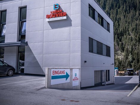 Ein modernes Gebäude mit einer hellen Fassade und dem Schriftzug „Bouvier“. Im Hintergrund erheben sich grüne Berge unter einem teilweise bewölkten Himmel.