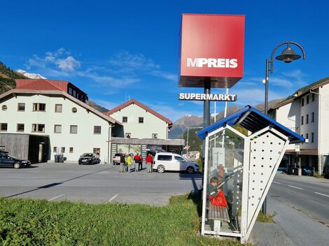 Ein Supermarkt mit dem Namen "MPREIS" ist in einer ländlichen Umgebung sichtbar. Im Vordergrund steht eine Bushaltestelle, und im Hintergrund sind mehrere Häuser zu sehen.