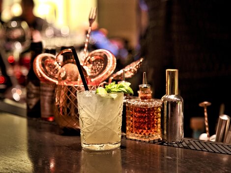 Ein leckerer Cocktail mit frischer Minze steht auf einer eleganten Bar. Im Hintergrund sind verschiedene Flaschen und Utensilien zu sehen.