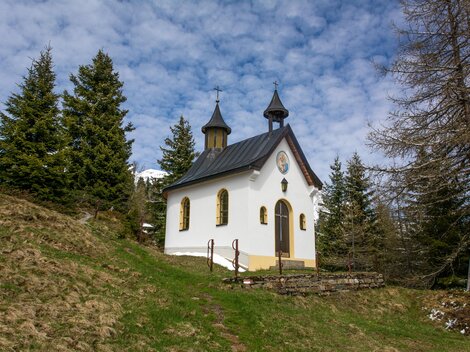 Eine kleine, charmante Kapelle steht auf einem Hügel, umgeben von grünen Wiesen und hohen Tannen. Der Himmel ist klar und zeigt einige Wolken. | © Bernhard Gruber – Photography