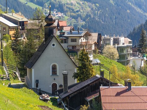 Ein malerisches Dorf in den Bergen mit einer kleinen Kirche. Die Umgebung ist grün und von beeindruckenden Hügeln umgeben. | © Bernhard Gruber – Photography