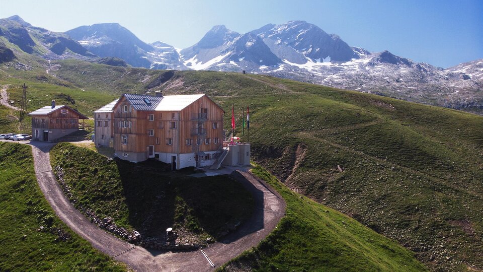 A modern building on a green hill, surrounded by mountains. In the background are snow-capped peaks and a clear blue sky.