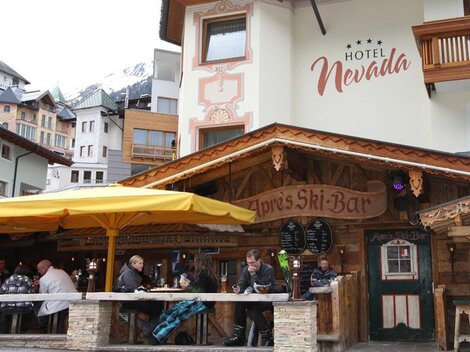 Ein gemütliches Restaurant mit einer Holzterrasse und einem großen Sonnenschirm. Menschen sitzen zusammen und genießen Getränke, während die Berge im Hintergrund sichtbar sind.