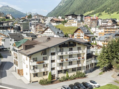 Ein malerisches Alpendorf mit typischen Chalets und einer schönen Berglandschaft. Im Vordergrund befindet sich ein modernes Gebäude mit Parkplätzen.