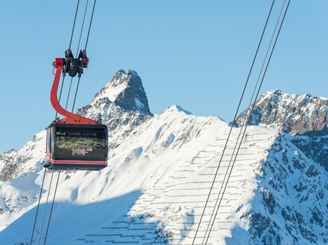 Eine Seilbahn schwebt über schneebedeckte Berge. Der Himmel ist klar und blau.