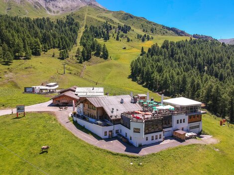 Ein gemütliches Bauernhaus inmitten einer grünen Berglandschaft. Im Hintergrund sind bewaldete Hänge und ein blauer Himmel zu sehen.