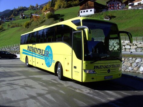 Ein gelber Reisebus steht an einem Parkplatz in einer bergigen Landschaft. Im Hintergrund sind grüne Hügel und einige Häuser zu sehen.