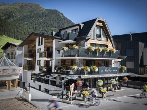 Ein modernes Hotel mit blühenden Balkonpflanzen in einer Berglandschaft. Die Fassade ist freundlich und einladend.