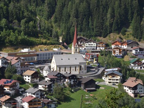 Eine malerische Bergstadt mit traditionellen Häusern und einer markanten Kirche. Umgeben von grünen Wiesen und dichten Wäldern.