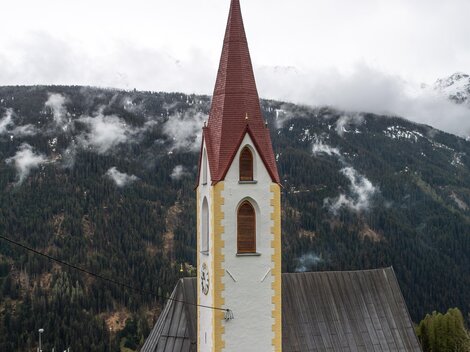 Eine Kirche mit einem roten Dach und einem hohen Kirchturm vor einer bergigen Landschaft. Die Umgebung ist von grünen Wäldern und sanften Hügeln geprägt. | © Bernhard Gruber – Photography