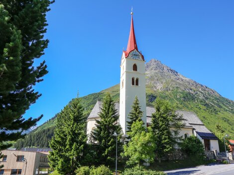 Eine malerische Kirche mit einem roten Turm, umgeben von grünen Bäumen und Bergen. Der Himmel ist klar und blau. | © TVB Paznaun - Ischgl
