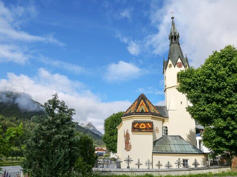 Eine malerische Kirche mit einem hohen Turm und buntem Dach. Im Hintergrund sind grüne Berge und ein blauer Himmel zu sehen.