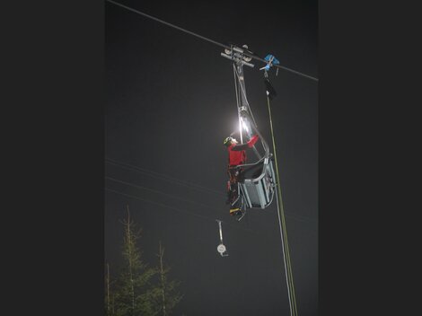 Eine Person arbeitet an einer Seilbahn in der Nacht. Sie trägt eine Sicherheitsausrüstung und nutzt eine Taschenlampe.