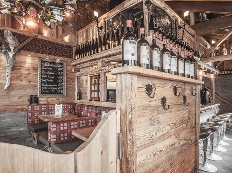 Ein gemütliches Restaurant mit Holzdekor und einer Bar. Die Flaschen stehen auf einem Holzregal, und es gibt Sitzgelegenheiten für Gäste.