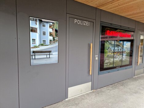 Ein moderner Polizeigebäudeeingang mit einem großen Fenster. Neben dem Eingang ist ein Bild einer Bank und Wohngebäude zu sehen.