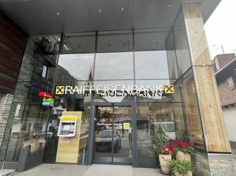 Ein modernes Bankgebäude mit großen Glasfenstern. Der Eingang gehört zur Raiffeisenbank und ist mit Pflanzen dekoriert.