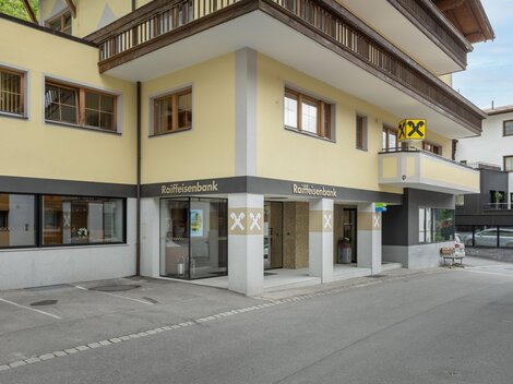 Ein modernes Bankgebäude mit gelber Fassade und großen Fenstern. Im Vordergrund ist ein Parkplatz und eine Straße sichtbar.