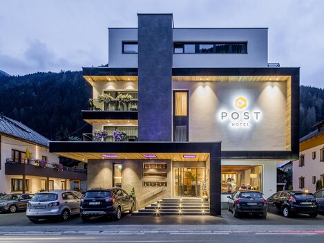 A modern hotel building with the inscription "POST" glows in the twilight. Surrounded by mountains and other buildings, it is an inviting place to stay.