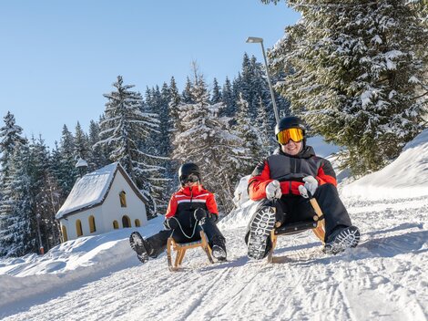 Eine verschneite Landschaft mit zwei Personen, die auf Schlitten fahren. Im Hintergrund sind winterliche Bäume und ein kleines, gemütliches Haus zu sehen.